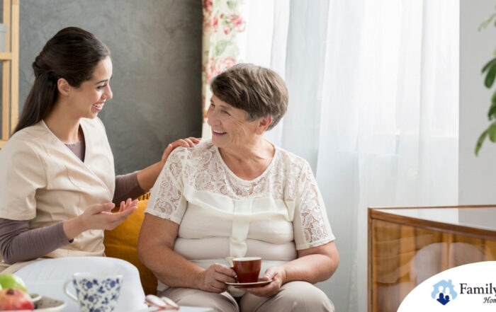 Smiling caregiver with her hand on an older woman’s shoulder as they share tea on the couch, showing home care vs assisted living