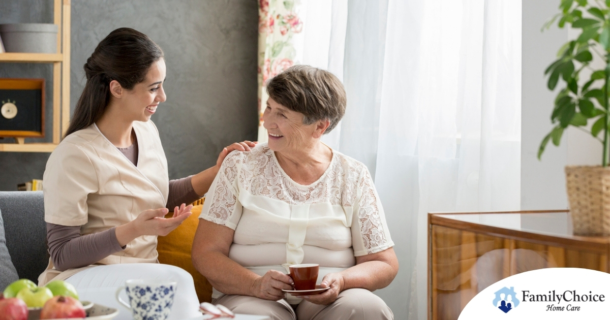 Smiling caregiver with her hand on an older woman’s shoulder as they share tea on the couch, showing home care vs assisted living